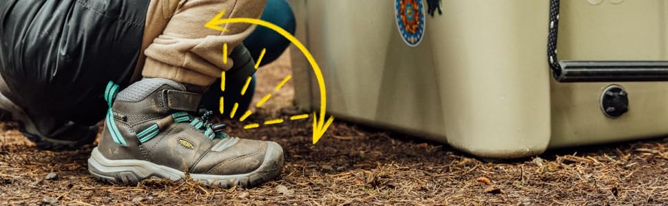 A child wears grey hiking boots with teal accents and KEEN.Bellows Flex in the foretoe kneeling by